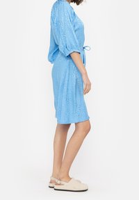 Jasnoniebieska sukienka z krótkim rękawem o teksturowanym, błyszczącym wykończeniu, ściśnięta w talii za pomocą wiązania. Zestawiona z beżowymi butami typu slip-on.