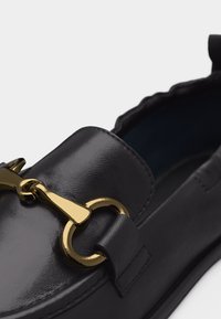 Mocassin en cuir noir avec détail en métal doré en forme de mors de cheval sur le dessus, coutures visibles et finition lisse et texturée.