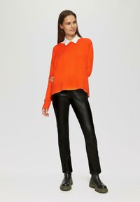 Leuchtend orangefarbener Strickpullover mit einem weißen Hemd darunter, kombiniert mit schwarzer Lederhose und schwarzen klobigen Stiefeletten.