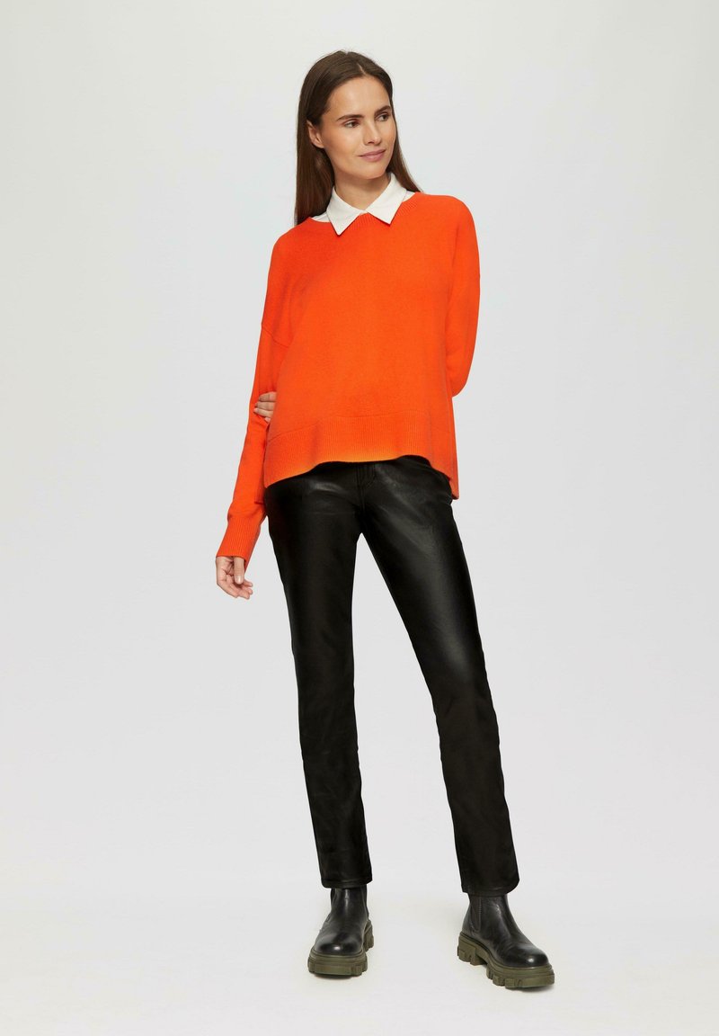 Leuchtend orangefarbener Strickpullover mit einem weißen Hemd darunter, kombiniert mit schwarzer Lederhose und schwarzen klobigen Stiefeletten.