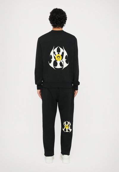 Sudadera negra y pantalones de chándal con gráficos de llamas blancas audaces y una cara sonriente amarilla en la parte trasera y en el muslo derecho, respectivamente.