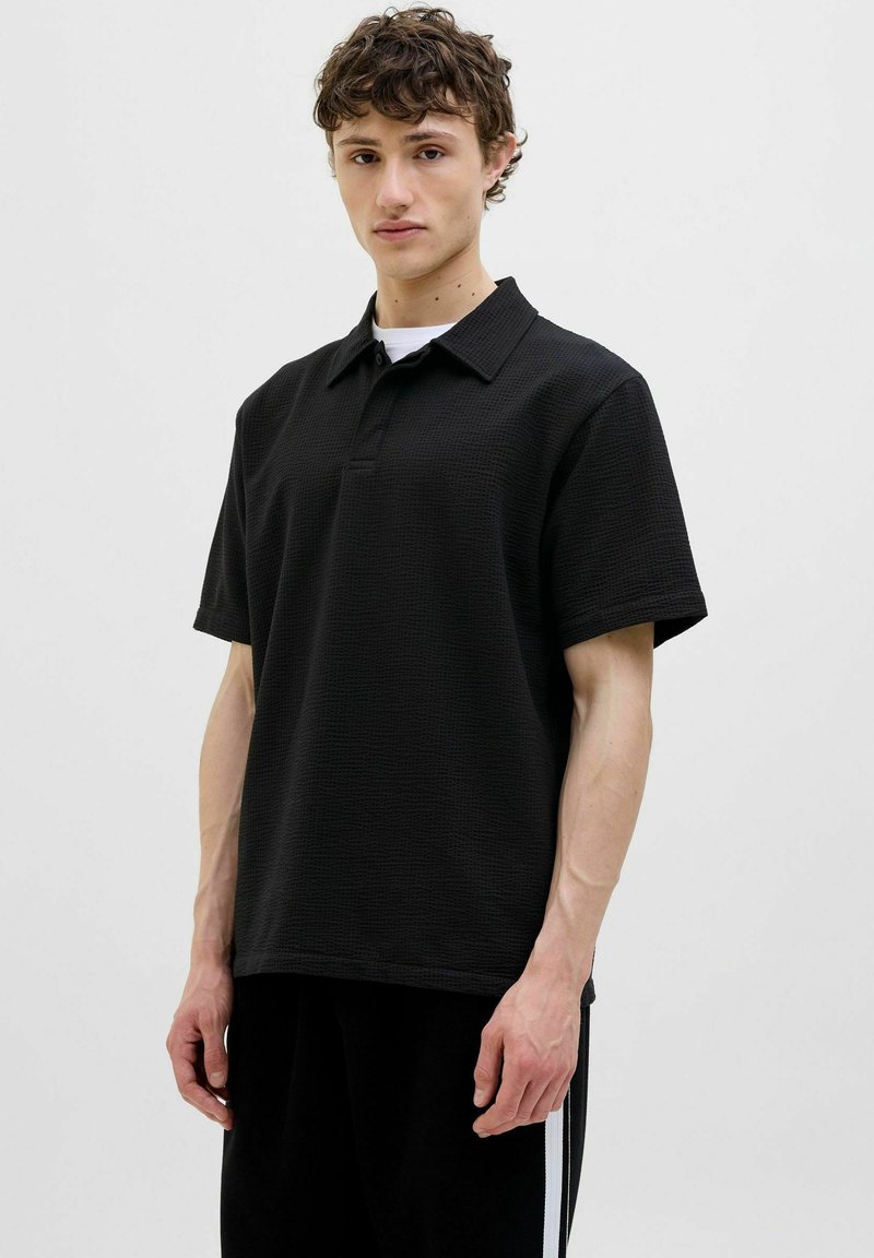 Sort polo-shirt med korte ærmer, lavet af tekstureret stof, knaplukning og klassisk krave. Modellen, der bærer den, står foran en ensfarvet baggrund.