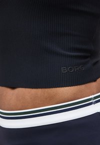 Κοντινό πλάνο στη μέση ενός ατόμου που φοράει μαύρο ριμπ τοπ με το κείμενο «BORG» και σκούρα μπλε παντελόνι με μέση με ρίγες σε λευκό, πράσινο και σκούρο μπλε.