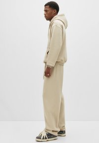Beige katoenen hoodie en bijpassende sweatpants, voorzien van een relaxte pasvorm, kangoeroezak en ribgebreide manchetten. Gecombineerd met donkere sneakers.