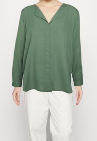 Blusa verde a maniche lunghe con scollatura a V e una texture morbida e leggera. Presenta una chiusura con bottoni e un orlo curvo.