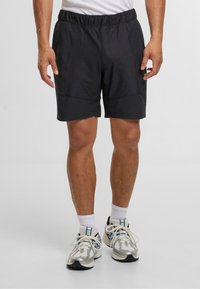Svarte sportsshorts med elastisk midje, med sidelommer og en lett buet bunnskjæring. Brukt med hvite sokker og sølvfargede joggesko.