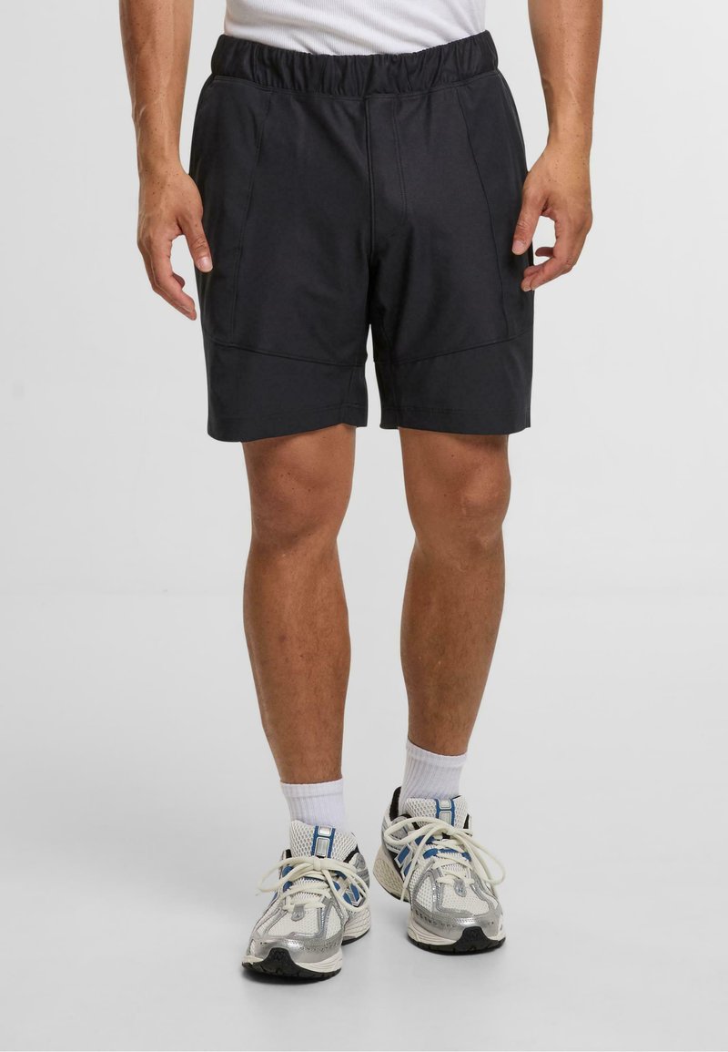Svarte sportsshorts med elastisk midje, med sidelommer og en lett buet bunnskjæring. Brukt med hvite sokker og sølvfargede joggesko.