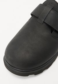 Zapato de cuero negro con puntera redondeada, cierre de correa y superficie texturizada. Presenta una suela resistente con un ligero patrón de relieve.