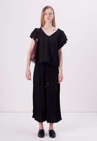 Black top with ruffled sleeves and textured fabric, paired with a long, flowing black skirt. Model holds a burgundy bag and wears black shoes.