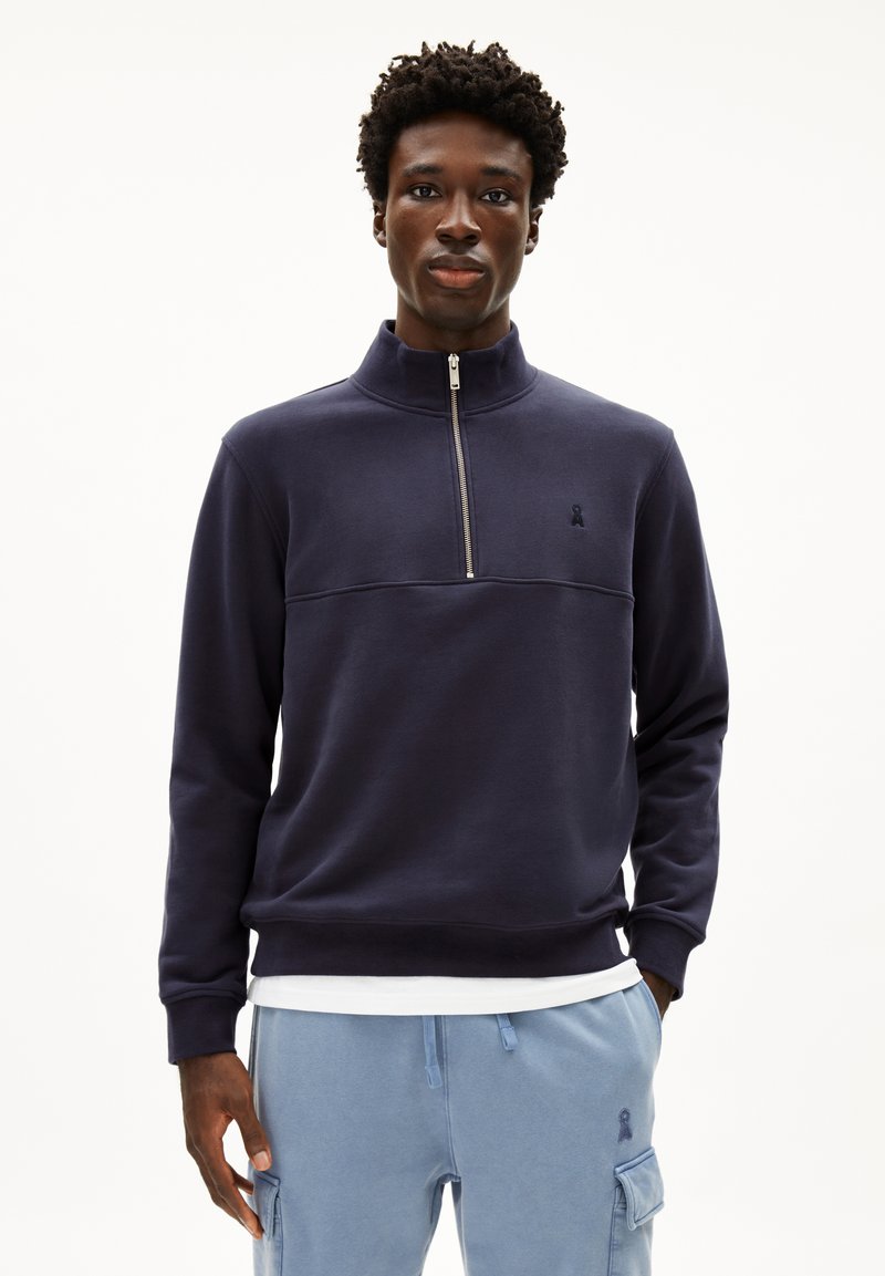 Sweat à col demi-zip en coton marine, avec un panneau sur la poitrine et un petit logo sur le côté gauche. Porté avec un pantalon bleu clair.