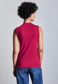 Tiivistetty hihaton paita kirkkaassa vaaleanpunaisessa värissä, valmistettu sileästä kankaasta, jossa on korkea kaula-aukko ja siisti muotoilu, yhdistetty sinisiin farkkuihin.