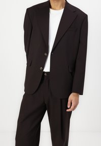 Ruskea pikkutakki, jossa on notchalapelli, kaksi nappia, sivutaskut ja neliömäinen taskutiedosto. Yhdistetty yhteensopiviin housuihin ja valkoiseen t-paitaan.