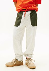 Pantalones joggers gris claro con acentos de panel verde oscuro, cinturilla elástica con cordón rojo y puños ajustados. Combinados con zapatillas beige.