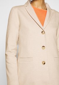Blazer beige con motivo testurizzato, con rever a lancia, tre bottoni e tasche laterali, indossato sopra una camicia arancione.