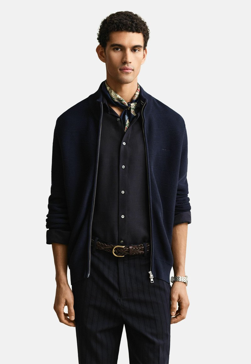 Homme portant un cardigan zippé bleu marine sur une chemise noire à boutons, une écharpe à motifs autour du cou, un pantalon rayé noir, une ceinture tressée et une montre argentée.