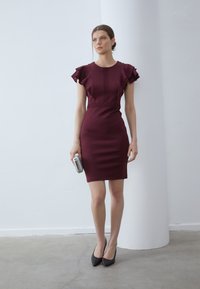 Burgundy klänning med åtsittande passform och volangärmar, spetsdetaljer ned längs framsidan, kombinerad med en silvrig clutch och svarta pumps med spetsig tå.