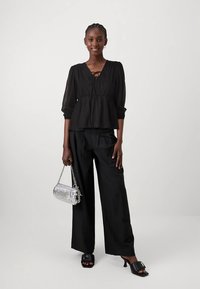 Kvinne iført en svart skjør bluse, vide sorte bukser, svarte hælede sandaler, og holder en liten sølvkjede håndveske.