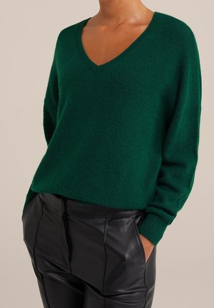 Personne portant un pull vert à col en V et un pantalon en cuir noir, les mains dans les poches, sur un fond uni.