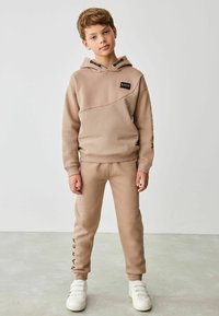 Béžová mikina s diagonálnym dizajnom, ktorá má malé čierne logo. Zodpovedajúce béžové joggers s nápisom "BAKER" v čiernom písme na nohe. Biely sneakers.