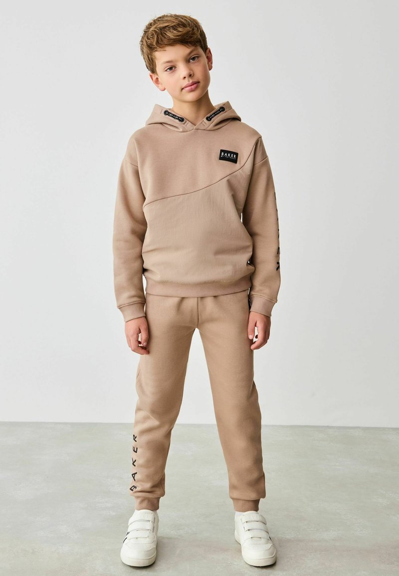 Béžová mikina s diagonálnym dizajnom, ktorá má malé čierne logo. Zodpovedajúce béžové joggers s nápisom "BAKER" v čiernom písme na nohe. Biely sneakers.