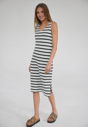 Jonge vrouw die glimlacht, draagt een mouwloze zwart-wit gestreepte midi-jurk met zijslitsen en bruine sandalen met gesp, staand op een effen achtergrond.