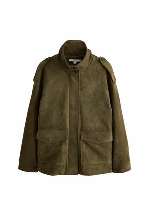 Kvinde med mørkt hår bundet tilbage iført en olivengrøn ruskindjakke over en beige skjorte og hvide bukser mod en beige baggrund.