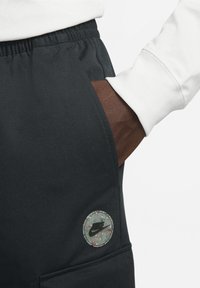 Joggers atléticos negros con una cintura elástica. Presentan un parche circular de Nike con un diseño moteado en el bolsillo lateral.