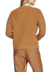 Brauner Strickpullover mit gerippter Textur, Rundhalsausschnitt und dezentem Knopfdetail am Rücken, kombiniert mit hellbraunen Lederhosen.