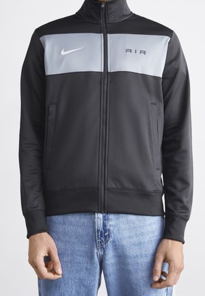 Černá bunda na zapínání na zip s šedým panelem na hrudi, bílý znak Nike vlevo, nápis "AIR" vpravo, nošena s světle modrými džíny.