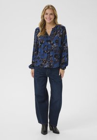 Donna sorridente con lunghi capelli biondi, che indossa una camicetta blu a fiori con bottoni, jeans a zampa scura e stivaletti neri, in piedi su uno sfondo bianco.