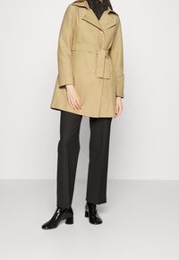 Beżowy trencz o podwójnym zapięciu, z paskiem w talii i szerokimi klapami. W zestawieniu z czarnymi spodniami i błyszczącymi botkami do kostek.