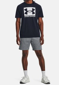 Marinblå t-shirt med en stor grå Under Armour-logga, i kombination med grå shorts. Modellen har på sig vita träningsskor och ankellånga strumpor.