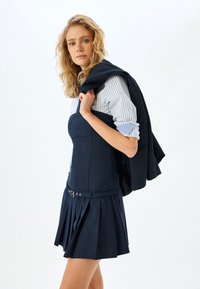 Marineblaues ärmelloses Kleid mit einem plissierten Rock und einem Gürtel; kombiniert mit einem hellblau gestreiften Hemd und einer dunklen Jacke, die über die Schulter gelegt ist.
