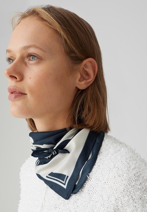 Vrouw met kort lichtbruin haar, gekleed in een wit textuurachtig shirtje en een zwart-wit zijden nekkruik met een patroon, vastgeknoopt in een klein knoopje.