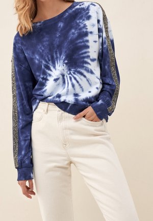 Kobieta ubrana w granatowo-biały sweatshirt z efektem tie-dye, z czarno-białymi paskami na rękawach oraz w kremowe, wysokowcieniowe spodnie.