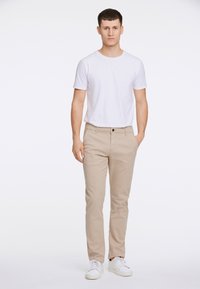 Beige katoenen broek met een rechte pasvorm, gecombineerd met een eenvoudig wit T-shirt. Het model staat zelfverzekerd in witte sneakers met de handen in de zakken.