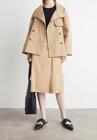 Beżowa podwójnie zapięta marynarka z dużym kołnierzem, zestawiona z pasującą spódnicą do kolan. Czarne płaskie buty i czarna torba na ramię dopełniają stylizację.