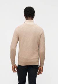 Άνδρας με κοντά σγουρά μαλλιά, ντυμένος με μπεζ πλεκτό πουλόβερ και μαύρα τζιν, στέκεται μπροστά σε απλό λευκό φόντο.