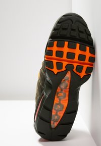 Podešev černé a oranžové tenisky Nike Air s texturovaným vzorkem, stojící vzpřímeně proti bílé stěně a povrchu.