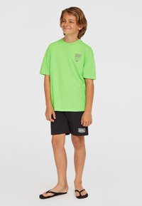 Camiseta verde brillante de manga corta, pantalones cortos negros informales y chanclas negras, con un pequeño logo en la camiseta y en los pantalones cortos.