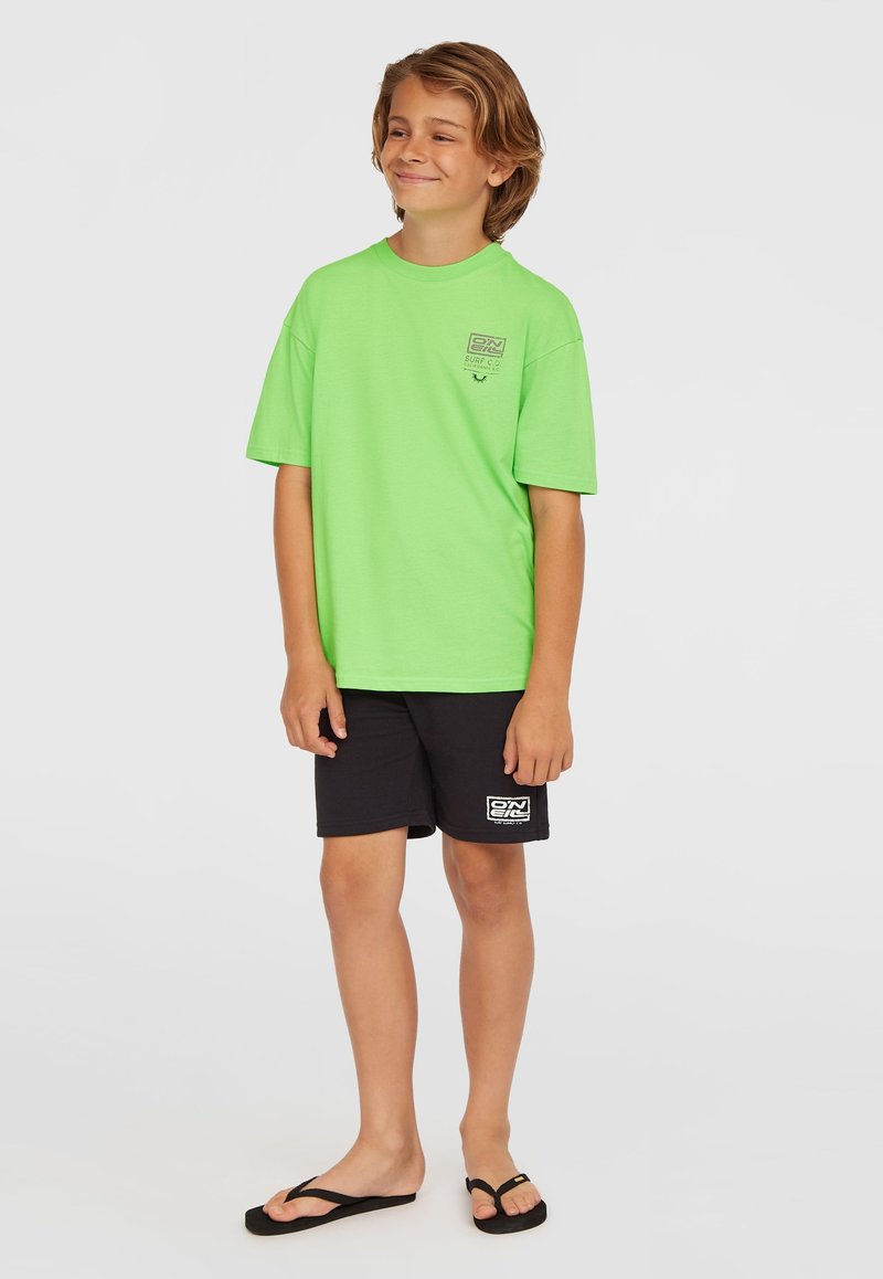 Camiseta verde brillante de manga corta, pantalones cortos negros informales y chanclas negras, con un pequeño logo en la camiseta y en los pantalones cortos.