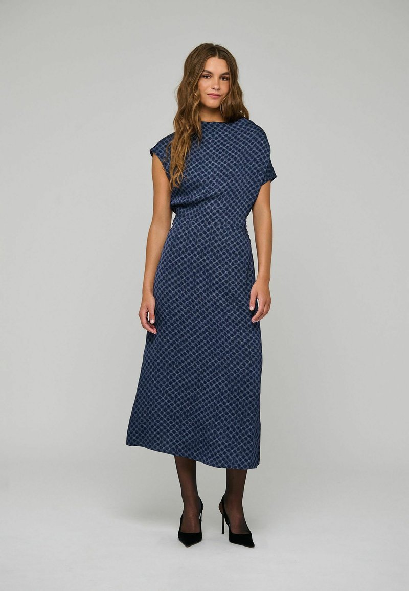 Femme debout portant une robe midi bleue à motifs avec des manches courtes, des collants noirs transparents et des chaussures à talons hauts noires sur un fond uni.