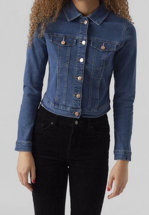Kvinde iført en tætsiddende blå denimjakke med knaplukning og sorte bukser, stående mod en ensfarvet baggrund.