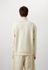 Sweatshirt crème à col montant, texture douce et coupe ample. Associé à un pantalon beige taille haute, à la fois minimaliste et décontracté.