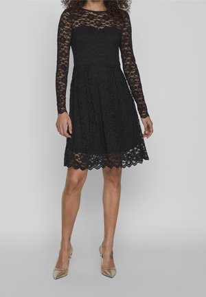 Black lace dress with long sleeves, round neckline, and scalloped hemline. Flared skirt and fitted bodice in textured lace fabric.