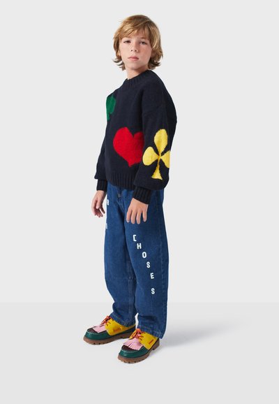 Pull en tricot navy avec des motifs verts, rouges et jaunes, associé à un jean bleu avec un texte blanc et des chaussures multicolores avec des accents contrastants.
