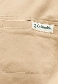 Τσέπη από μπεζ ύφασμα με ραμμένο άκρο και μικρή λευκή ετικέτα που εμφανίζει το πράσινο λογότυπο της Columbia Sportswear Company.