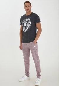 Graues T-Shirt mit weißem Grafikdruck, der Palmen zeigt, kombiniert mit roten melierten Jogginghosen und weißen Turnschuhen. Einfaches, lässiges Design.