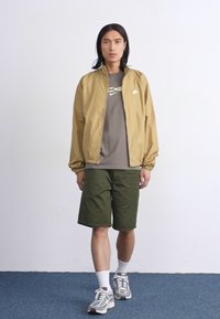 En ung mann med langt hår har på seg en beige jakke, grå skjorte og grønne shorts, og står på et blått teppe mot en enkel hvit vegg.