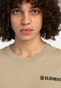 Camiseta de manga corta beige con cuello redondo y el logo negro "ELEMENT" en el pecho. Tejido suave con una textura lisa.